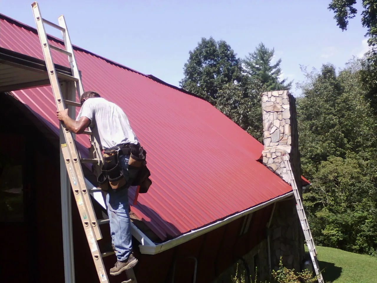 Roofer on a ladder beside a red metal roof and stone chimney — Soffit & Fascia Repair by Roofing Brothers in Swayzee